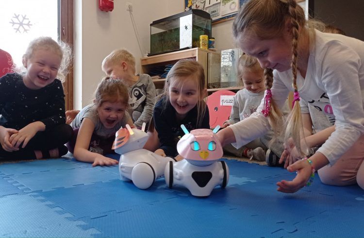 Grupa roześmianych dzieci klęczy, siedzi lub półleży na macie, bawiąc się robotami do programowania