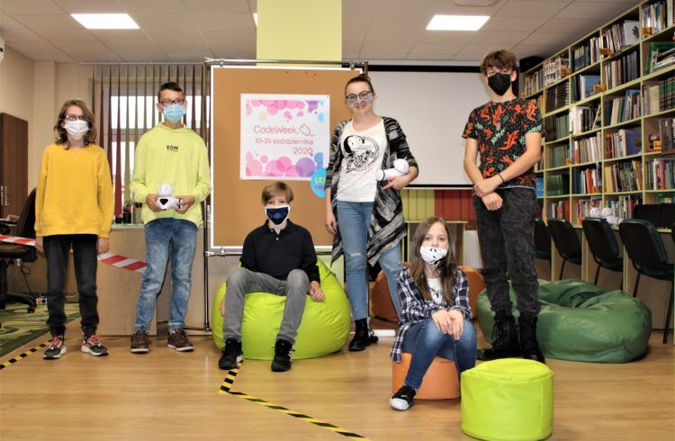Na fotografii grupa dzieci biorących udział w zajęciach programowania robotów wraz z bibliotekarką. Dwie osoby trzymają w rękach roboty.
