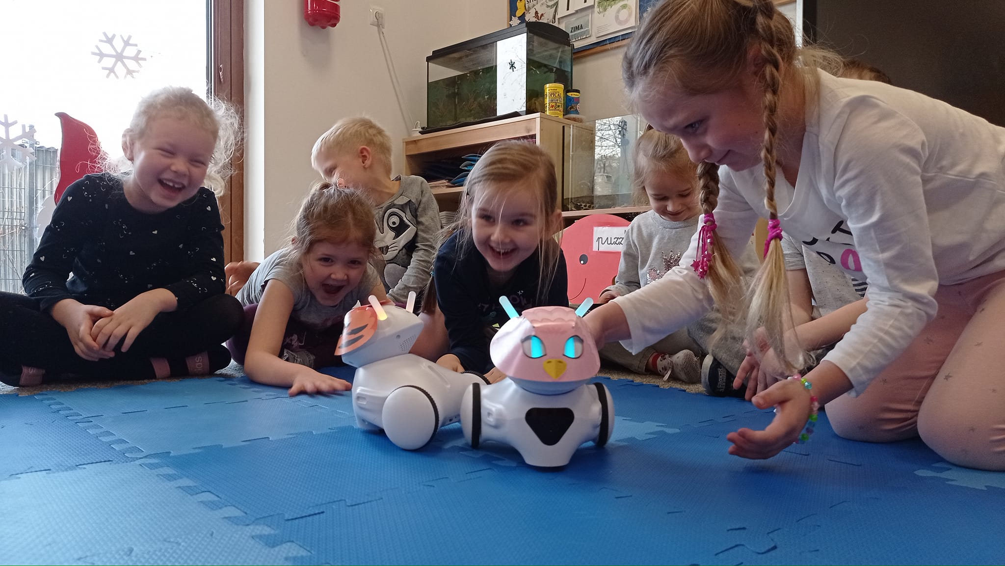 Grupa roześmianych dzieci klęczy, siedzi lub półleży na macie, bawiąc się robotami do programowania
